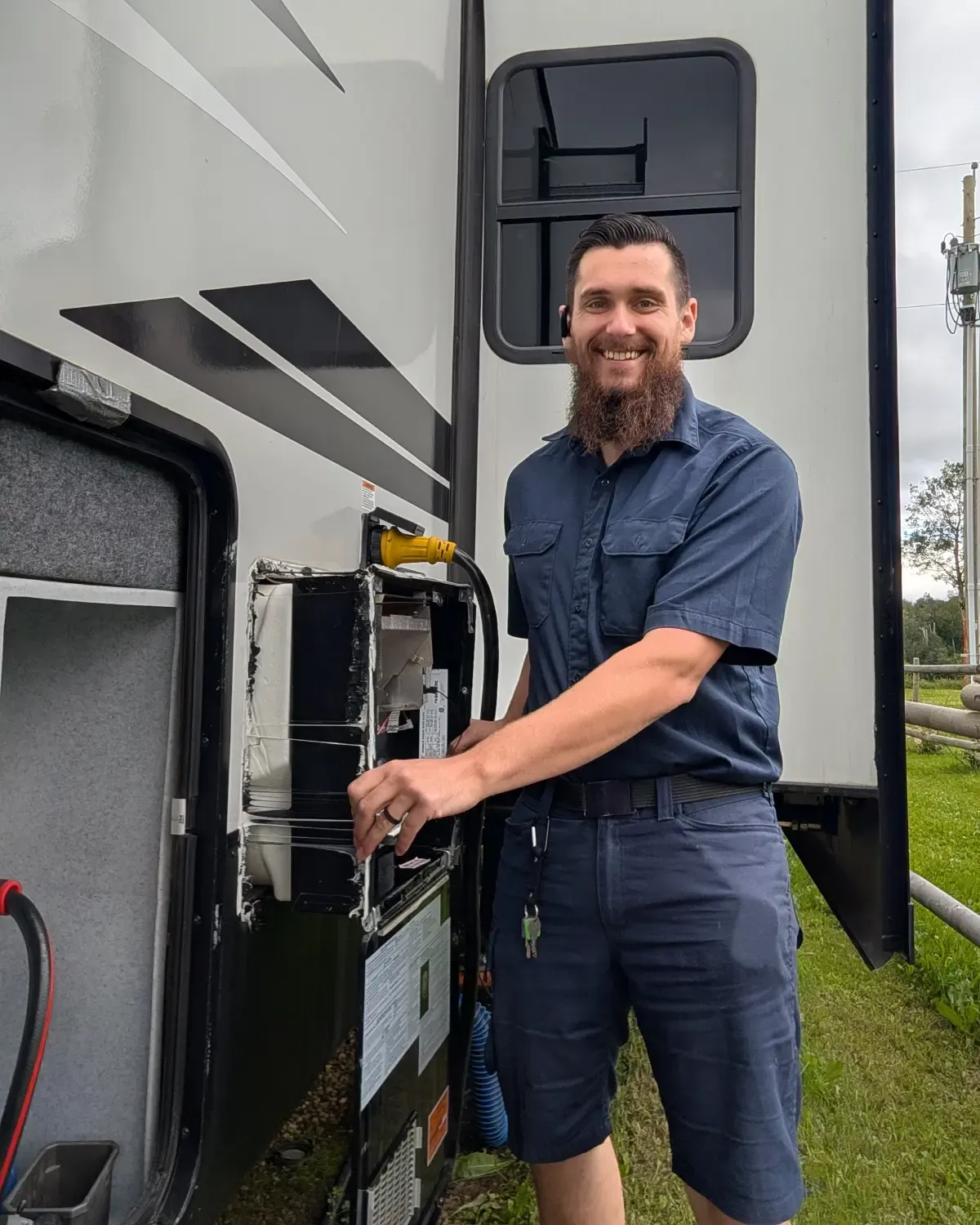 Kolton White, Red Seal RV technician, working on the open exterior appliance bay of a travel trailer parked on grass.