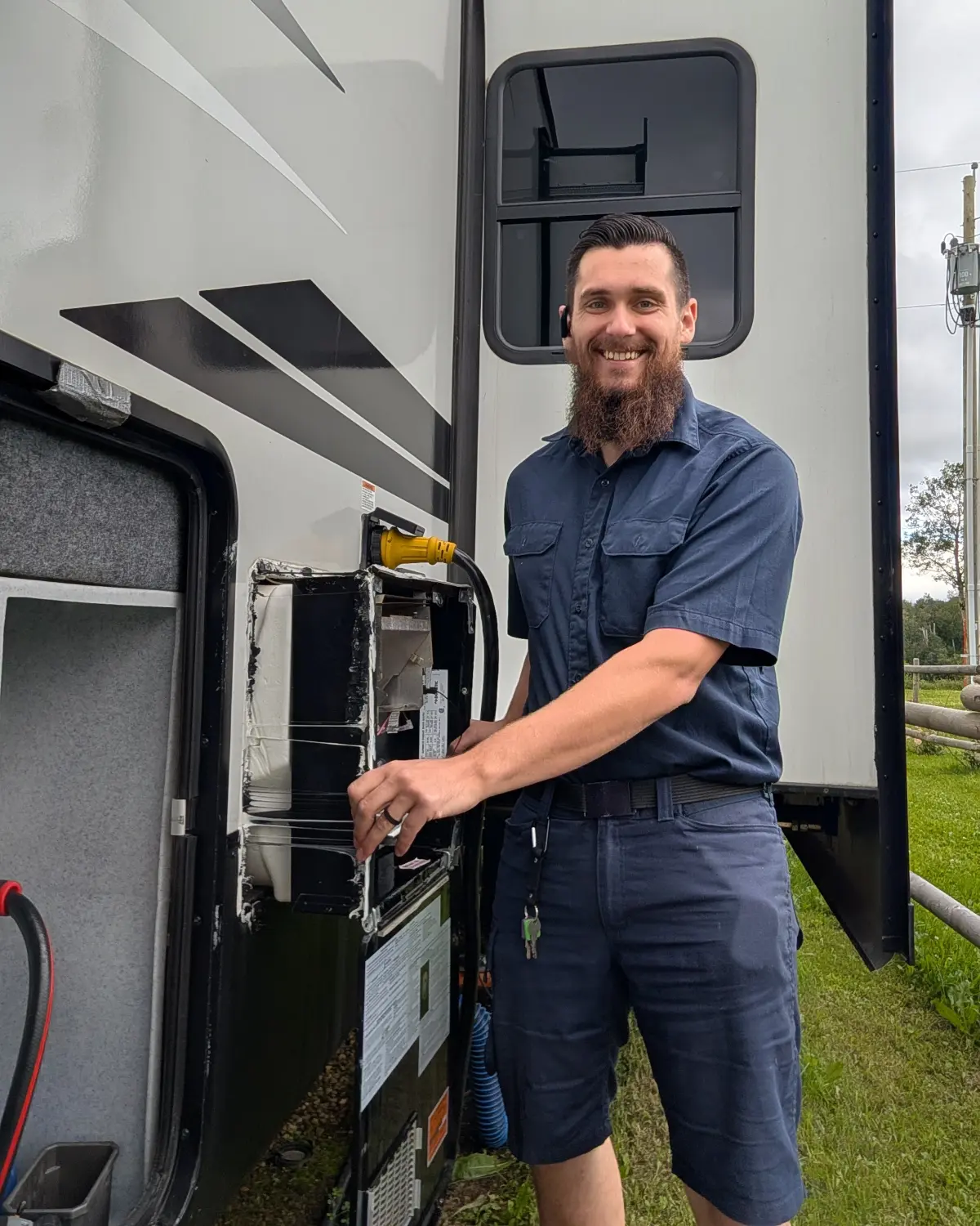 Kolton White, Red Seal RV technician, working on the open exterior appliance bay of a travel trailer parked on grass.